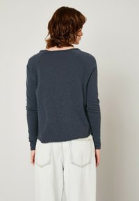 Tamsiai mėlynos spalvos ilgomis rankovėmis marškiniai su laisvu siluetu ir tekstūruota medžiaga. Gysliniai rankogaliai ir šiek tiek trumpesnis apdailos kraštas. Dėvima su šviesios spalvos plačiąja kelnėmis.