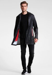 Άντρας περπατάει φορώντας μαύρο μακρύ αδιάβροχο με κόκκινη και λευκή επένδυση, μαύρο πουκάμισο, στενά τζιν και μαύρα αθλητικά.