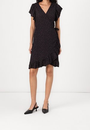 Femme portant une robe portefeuille noire avec un petit imprimé floral rose, manches volantées, ourlet à volants et escarpins slingback noirs à bout pointu.