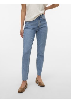 Femme portant un jean slim taille haute bleu clair avec ourlet brut et des chaussures beige à petit talon, debout devant un fond blanc uni.