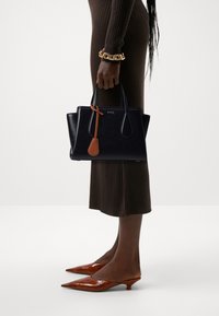 Svart läderhandväska med rundade kanter, orange accenttagg och strukturerad form. Modellen bär en ribbad brun klänning och spetsiga bruna klackar.