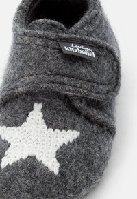 Pantofole grigie con un design a stella bianca lavorata a maglia, caratterizzate da una morbida texture e un'etichetta nera con il nome del marchio.