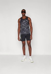 Débardeur de sport noir avec un motif tourbillonnant, associé à un short noir. La tenue comporte un logo sur le haut et des accents latéraux. Baskets blanches.