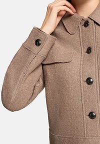 Brązowy wełniany płaszcz z złożonym kołnierzem, dużymi czarnymi guzikami i przednią kieszenią. Materiał ma teksturowane, lekko puszyste wykończenie.