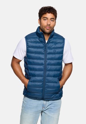 Chaleco acolchado azul marino con cuello alto, cierre de cremallera y bolsillos laterales. Hecho de material sintético suave. El usuario tiene las manos en los bolsillos.