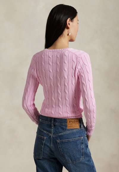 Frau mit langen dunklen Haaren, die einen rosa Strickpullover mit Zopfmuster und blaue Jeans mit Polo-Label trägt, mit den Händen in den Taschen stehend, von hinten betrachtet.