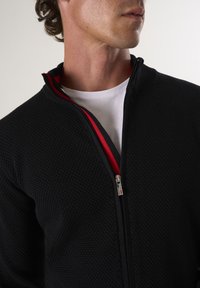 Giacca nera con zip e un motivo testurizzato, con un accento rosso all'interno del colletto e una zip argentata. Indossata sopra una t-shirt bianca.