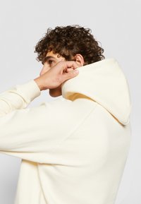 Hoodie crème avec une texture douce, capuche ajustable avec cordon de serrage et poignets côtelés. Présente une coupe décontractée et des coutures soignées.