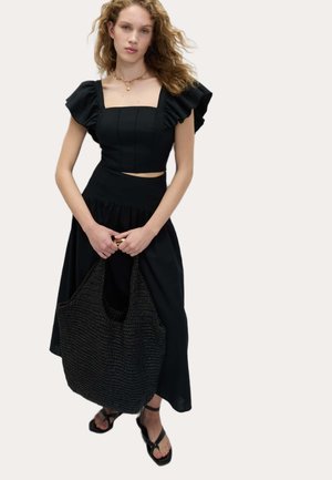 Sort to-delt outfit med voluminøse ærmer, tætsiddende top og flagrende nederdel. Holder en tekstureret sort vævet taske. Sorte flade sandaler.