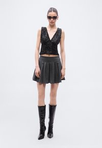 Μαύρο τοπ με παγιέτες και βαθύ V, συνδυασμένο με γκρι πλισέ μίνι φούστα και μαύρες μπότες από πατενέ δέρμα μέχρι το γόνατο.