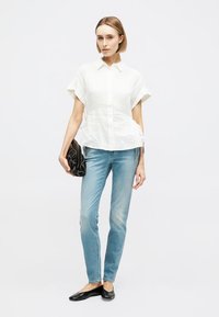 Femme portant une chemise blanche à manches courtes boutonnée, un jean skinny bleu clair, des ballerines noires, tenant une pochette noire, debout devant un fond blanc.