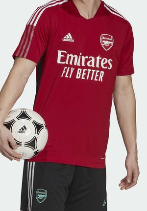 Person trägt ein rotes Arsenal-Fußballtrikot und schwarze Shorts, hält einen schwarz-weißen Adidas-Fußball in der linken Hand.