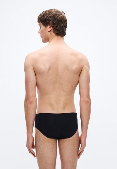 Schwarze Badehose aus glattem Material, figurbetontes Design, niedrige Taille und minimale Bedeckung, getragen von einer Person aus der Rückenansicht.