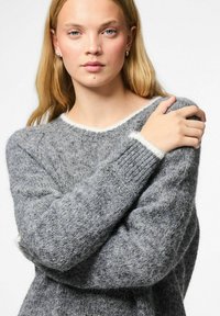 Giovane donna bionda con occhi chiari che indossa un maglione grigio morbido, che tiene la spalla sinistra con la mano destra su uno sfondo bianco semplice.