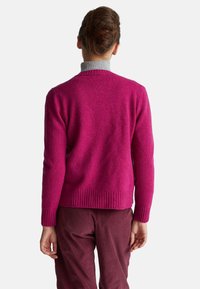 Světle fuchsiový svetr vyrobený z měkkého materiálu, s ribbovaným lemem a manžetami. Nosený s tmavě vino červenými kalhotami, zobrazeno zezadu.