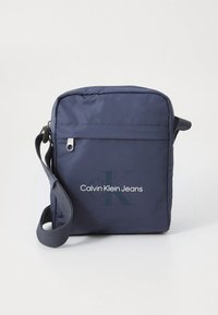 Laivastonsininen nylonista valmistettu olkalaukku, jossa säädettävä hihna, etutasku vetoketjulla ja valkoinen logo "Calvin Klein Jeans" isolla K-kirjaimella.