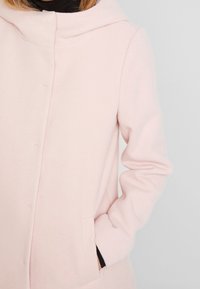 Cappotto rosa chiaro realizzato in tessuto morbido, con cappuccio, maniche lunghe e tasche laterali, caratterizzato da un semplice design con chiusura a bottoni.