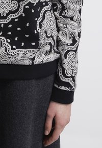 Svart sweatshirt med intrikata vita paisleymönster, med ribbade muddar och en bekväm passform. Tyget verkar mjukt och texturerat.