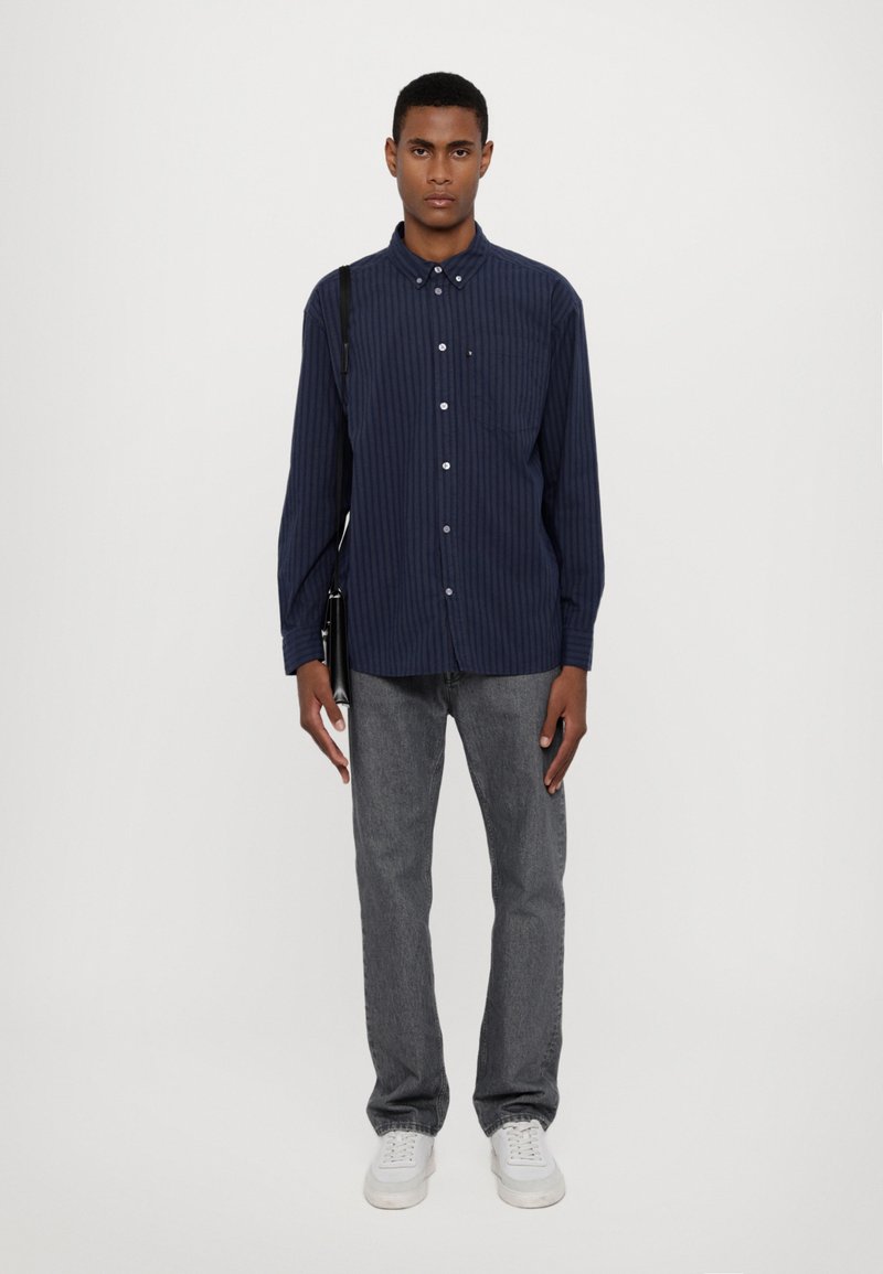 Marine gestreepte overhemd met lange mouwen, met knoopsluiting en borstzak, gecombineerd met grijze denim jeans en witte sneakers. Minimalistische tas op de schouder.