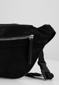 Marsupio in suede nero con forma curva, chiusura con zip argentata e tracolla regolabile in tessuto. Superficie texturizzata con accenti lucidi.