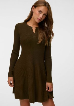 Femme aux longs cheveux bruns portant une robe marron foncé moulante à côtes, à manches longues, avec un décolleté cranté, debout devant un fond uni.