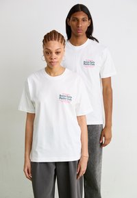 Two individuals wear oversized, short-sleeve white t-shirts. The shirts feature colorful text: "Buena Pasta Pesto Club" and a logo.