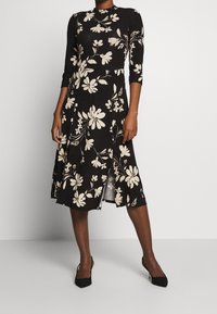 Czarna sukienka w kwiaty z długimi rękawami, z białymi kwiatami i rozcięciem z przodu. Wykonana z gładkiego materiału o dopasowanym kroju.