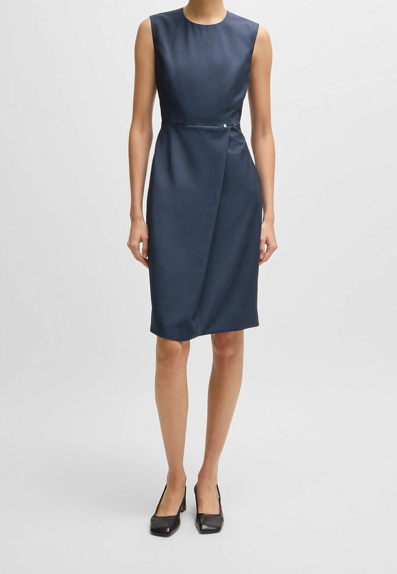Femme portant une robe portefeuille sans manches bleu marine longueur genou avec un petit bouton latéral, associée à des chaussures noires à talons bas.