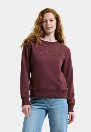 Woman with red hair wearing a maroon Didriksons sweatshirt and light blue jeans standing against a plain background.
