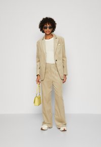 Beige corduroy blazer og bukser sæt, har et tætsiddende design med elastisk talje. Kombineret med en hvid top, en gul håndtaske og solbriller.
