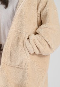 Beige fleecjack met een zachte textuur, voorzien van een voorkruiszak en ruime mouwen. De stof is pluizig en dik voor warmte.