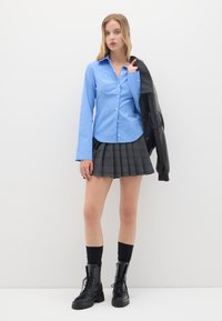 Camicia azzurra a righe con bottoni, gonna grigia a pieghe, calze nere fino al ginocchio e stivali militari. Giacca di pelle nera appoggiata su una spalla.