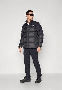 Muškarac u crnoj jakni na preklop, crnim hlačama, crnim tenisicama i velikim crnim sunčanim naočalama, stoji nasuprot jednostavne svijetle pozadine.