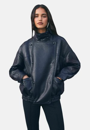 Femme portant une veste en cuir noire texturée oversize avec un col montant et les mains dans les poches, associée à un pantalon noir.