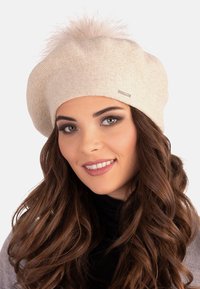 Beige kötött beret, pufók pom-pommal a tetején, sima textúrával és diszkrét fényességgel. Egy kis fém címkével díszítve.