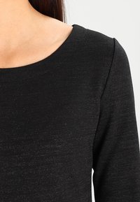 Czarna bluzka z dekoltem w łódkę. Tkanina ma subtelny połysk i teksturowany wygląd. Posiada długie rękawy i klasyczny krój.