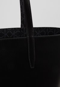 Černá taška přes rameno s texturovaným koženým povrchem a kontrastní vnitřní částí se subtilním logem. Pevné ploché uši pro snadné nošení.