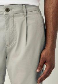 Lysgrå bukser lavet af glat stof, med frontknaplukning, sidelommer og en plisseret design langs benet.