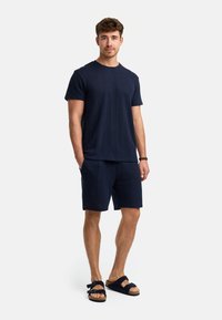 Marineblaues T-Shirt mit kurzen Ärmeln aus strukturiertem Stoff, kombiniert mit passenden Shorts. Getragen mit schwarzen Sandalen mit doppelten Riemen.