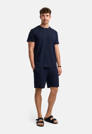 Marineblaues T-Shirt mit kurzen Ärmeln aus strukturiertem Stoff, kombiniert mit passenden Shorts. Getragen mit schwarzen Sandalen mit doppelten Riemen.