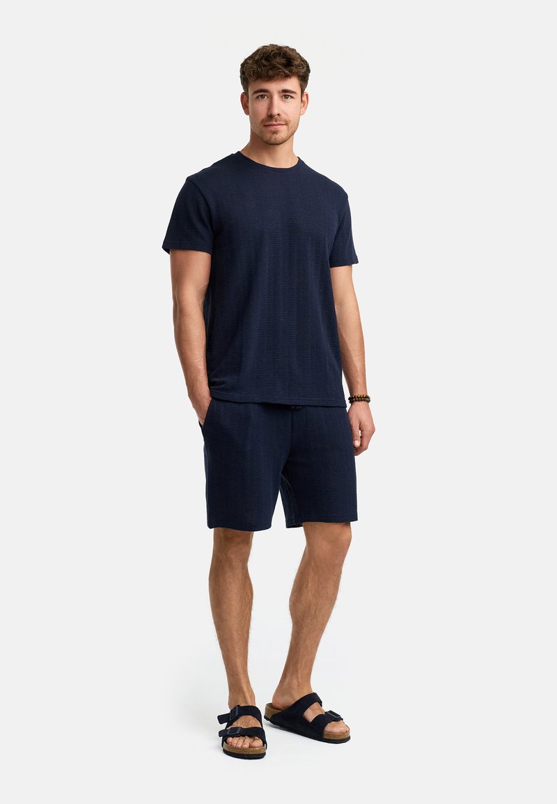 Marineblaues T-Shirt mit kurzen Ärmeln aus strukturiertem Stoff, kombiniert mit passenden Shorts. Getragen mit schwarzen Sandalen mit doppelten Riemen.