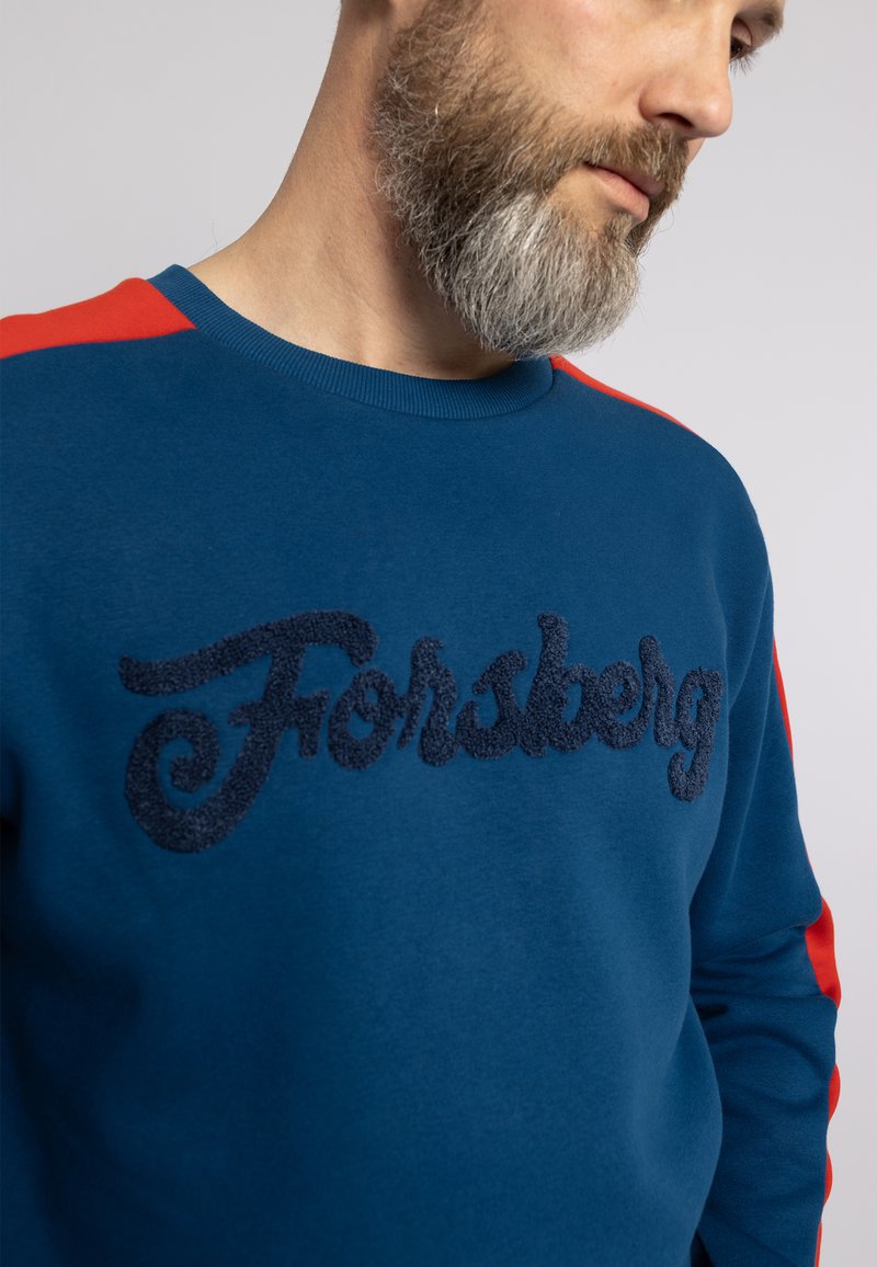 Sudadera azul con un logo bordado en relieve en navy "Forsberg", con acentos en rojo en los hombros y un cuello redondeado.