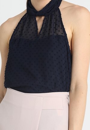 Femme portant un haut noir sans manches à col halter avec des pois texturés, associé à un pantalon taille haute rose pâle.