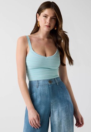 Femme aux longs cheveux bruns portant des boucles d'oreilles créoles dorées, un débardeur en maille texturée bleu clair et un jean large taille haute.