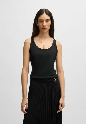 Musta ribbetty tank top, jossa on pyöreä kaula-aukko, yhdistettynä laskostettuun mustaan hameeseen. Hameen metalliset yksityiskohdat ja herkkä riipuskaulakoru.