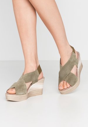 Pieds portant des sandales compensées en daim vert olive avec des brides croisées et un talon plateforme tissé sur un sol gris clair.