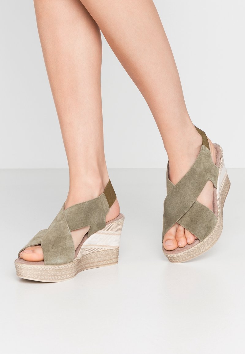 Pieds portant des sandales compensées en daim vert olive avec des brides croisées et un talon plateforme tissé sur un sol gris clair.