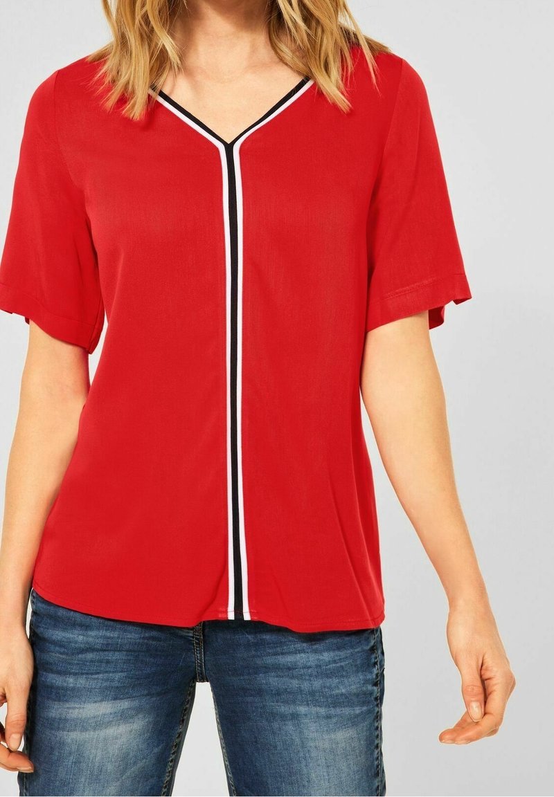 Femme portant un chemisier rouge à manches courtes avec un col en V et une rayure verticale noire et blanche au centre, associé à un jean bleu.