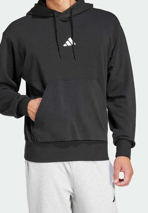 Man draagt een zwarte Adidas hoodie met voorzak en logo, gecombineerd met lichtgrijze joggingbroek.