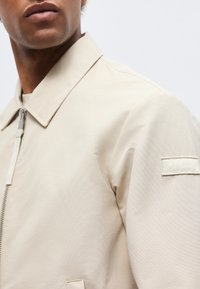 Mand iført en beige jakke med lynlås og krave, med en lille rektangulær lap på den øverste ærme og en sidelomme.